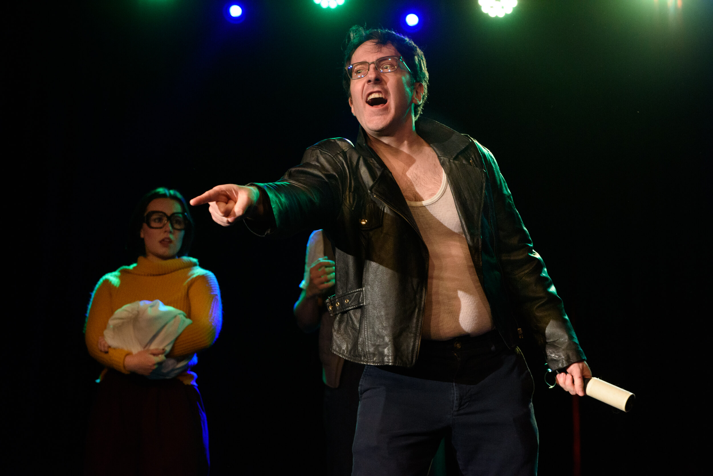 Taylor is wearing a black leather jacket and pointing to the audience with an angry look on his face. Behind him, Islay looks shocked.