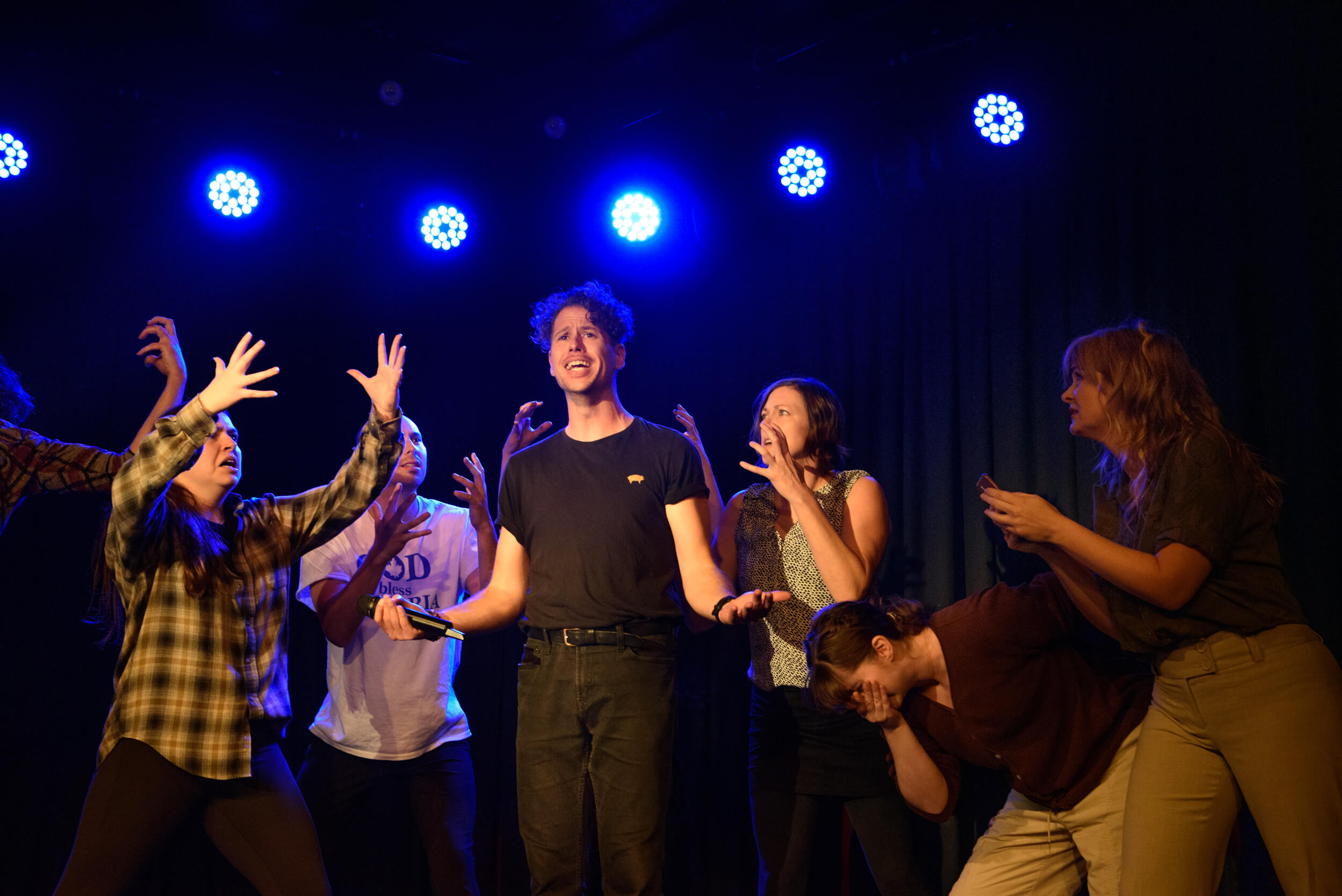 A group of performers are gathered around Tony - they have their hands in the air and look angry. Tony is holding out his hands with an uncomfortable expression on his face.